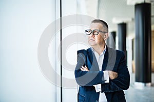 Seniow businessman with crossed hands standing and thinking by the panoramic windows in office.