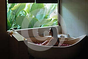 Sensitive young woman enjoying in spa. Luxury stone bath tub with jungle view near window. Natural organic tropical petals in the