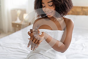 Sensual African American woman sitting on bed in bath towel, applying nourishing cream on her hands at home