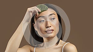 Sensual asian lady doing daily skin care routine, massaging facial skin with jade gua sha scrapper, brown background