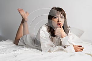 Sensual beautiful young woman wearing a white men& x27;s shirt lies on bed. Sexy model with finger on mouth looking at