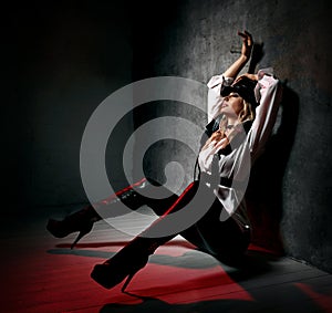 Sensual blonde woman in leather pants, white shirt and leather belts sits at concrete wall, hands up, feeling arousal