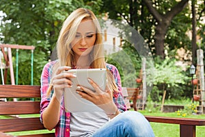 Sensual blonde woman sitting in park on wooden bench. She is using tablet pc. Outdoor photo. She looks relaxed.