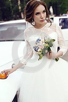 Sensual bride with dark hair in luxurious wedding dress posing beside car