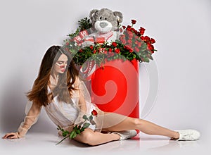 Sensual brunette woman in white stylish bodysuit and sneakers lying at huge box with flowers looking at rose in her hand