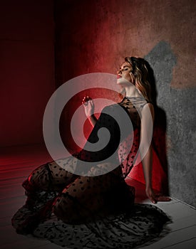 Sensual excited young blonde curly woman in transparent lace dress sits on knees on floor at wall with eyes closed