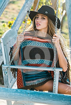 Sensual fashion woman in boho style swinging on swing on summer nature, young girl in hat relaxing in spring park