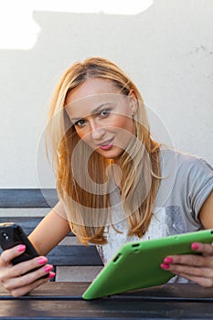 Sensual happy blonde woman sitting on wooden bench. She is using mobile phone and tablet pc. Outdoor photo. She looks relaxed