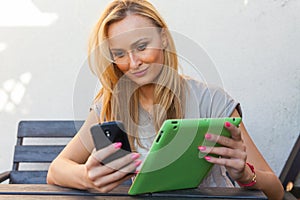 Sensual happy blonde woman sitting on wooden bench. She is using mobile phone and tablet pc. Outdoor photo. She looks relaxed