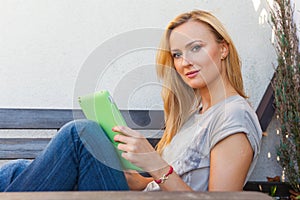 Sensual happy blonde woman sitting on wooden bench. She is using mobile tablet pc. Outdoor photo. She looks relaxed.