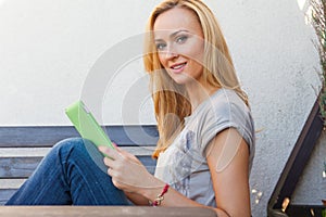 Sensual happy blonde woman sitting on wooden bench. She is using mobile tablet pc. Outdoor photo. She looks relaxed.