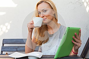 Sensual happy blonde woman sitting on wooden bench. She is using mobile tablet pc. Outdoor photo. She looks relaxed