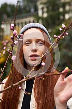 Sensual portrait of beautiful redhead woman in hat smelling pink flowers in spring