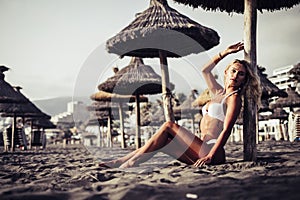 Sensual portrait of beautiful yhoung blonde female taking bathsun at the beach sit down on the sand with white bikini on the dark