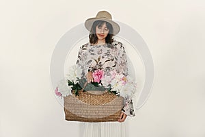 Sensual portrait of boho girl holding pink peonies in rustic basket, isolated on white. Stylish hipster woman in hat and bohemian
