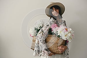 Sensual portrait of boho girl holding pink and white peonies in rustic basket. Stylish hipster woman in hat and bohemian floral