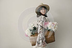 Sensual portrait of boho girl holding pink and white peonies in rustic basket. Stylish hipster woman in hat and bohemian floral