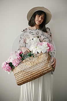 Sensual portrait of boho girl holding pink and white peonies in rustic basket. Stylish hipster woman in hat and bohemian floral