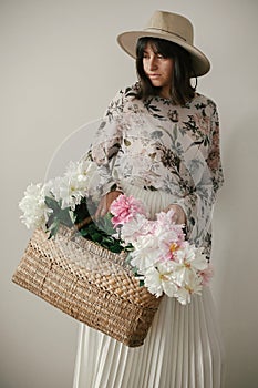 Sensual portrait of boho girl holding pink and white peonies in rustic basket. Stylish hipster woman in hat and bohemian floral