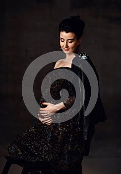 Sensual pregnant noble woman in black off-shoulder dress and leather jacket stands holding hand at her belly, feeling love