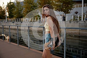 Sensual slim young woman walking with longboard over summer cityscape