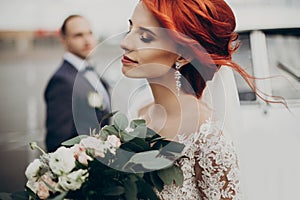 Sensual stylish bride holding modern bouquet and relaxing with windy hair and groom looking at her near retro car. luxury wedding