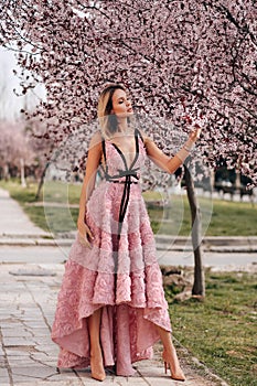 Sensual woman with blond hair in elegant dress and accessories posing among blossoming peach trees in park