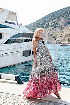 Sensual woman with blond hair in luxurious dress with accessories posing near yacht in dock