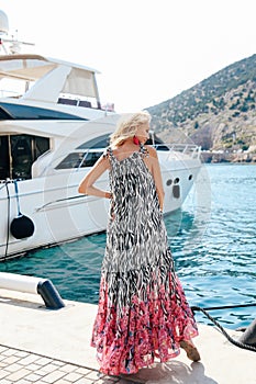 Sensual woman with blond hair in luxurious dress with accessories posing near yacht in dock