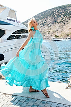 Sensual woman with blond hair in luxurious dress with accessories posing near yacht in dock