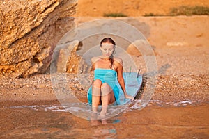 Sensual woman in blue dress sitting on surfboard at sea beach. Summer vibes, active lifestyle and beach vacations idea