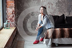 Sensual woman with dark hair in blue shirt and jeans sitting on a bed at home. Loft style interior