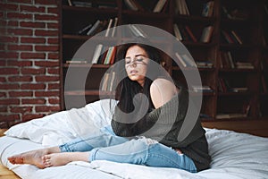 Sensual woman with dark hair in green sweater and jeans sitting on a bed