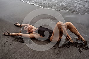 Sensual woman in fashionable black swimsuit lying on the black sand, on the beach, smiling, having fun