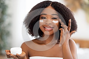 Sensual young black lady applying moisturizing facial cream from jar near mirror at home. Beauty and natural cosmetics