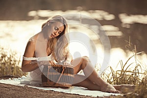 Sensual young woman on the picnic sitting by the lake at sunset and looking in picnic basket