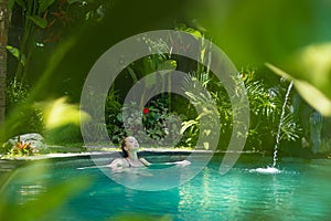 Sensual young woman relaxing in outdoor spa infinity swimming pool surrounded with lush tropical greenery of Ubud, Bali.