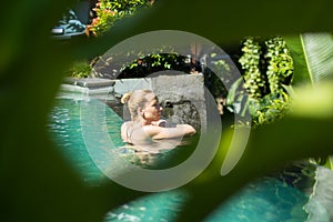 Sensual young woman relaxing in outdoor spa infinity swimming pool surrounded with lush tropical greenery of Ubud, Bali.