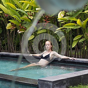 Sensual young woman relaxing in outdoor spa infinity swimming pool surrounded with lush tropical greenery of Ubud, Bali.