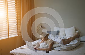 Sensual young woman in white tank top and panties lying on bed near laptop