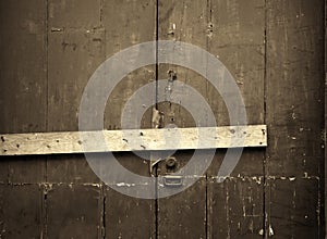 sepia close up of an old cracked peeling wooden plank door barred shut with a piece of timber and rusty nails with a lock and