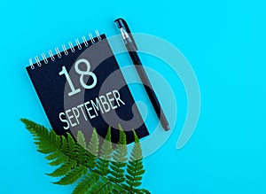 September 18th. Day 18 of month, Calendar date. Black notepad sheet, pen, fern twig, on a blue background