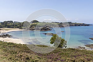 a serene beach scene with calm, turquoise waters, a sandy shore bordered by rocky outcrops and lush greenery under a clear sky. A