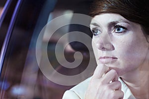 Serene businesswoman with hand on chin looking through car window at the city nightlife, reflected lights