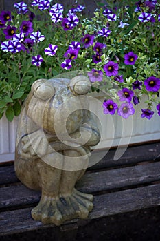 Serene concrete frog sitting on bench with purple flowers.