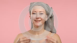 Serene female with towel on head gently applies hydrating facial mask on plain pink background