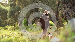 A serene lady in a hat with her loyal Corgi, both enjoying a serene walk through an olive grove