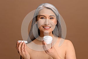 Serene middle-aged Asian woman with fresh clean healthy skin stands with jar of moisturiser anti-aging cream