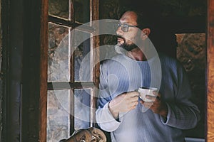 Serene and relaxed mature man standing indoor at home near the window and looking outside thinking. Real people lifestyle. Male