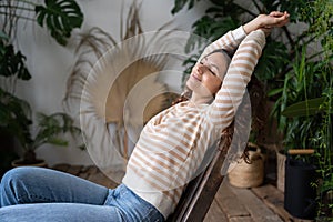 Serene relaxed young woman with closed eyes stretching arms, relaxing in urban jungle interior